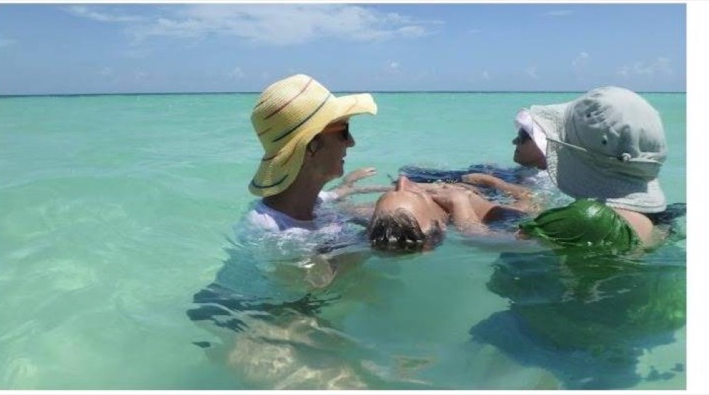Caroline and a colleague in wide-brimmed hats supporting a person floating on their back in shallow turquoise sea.