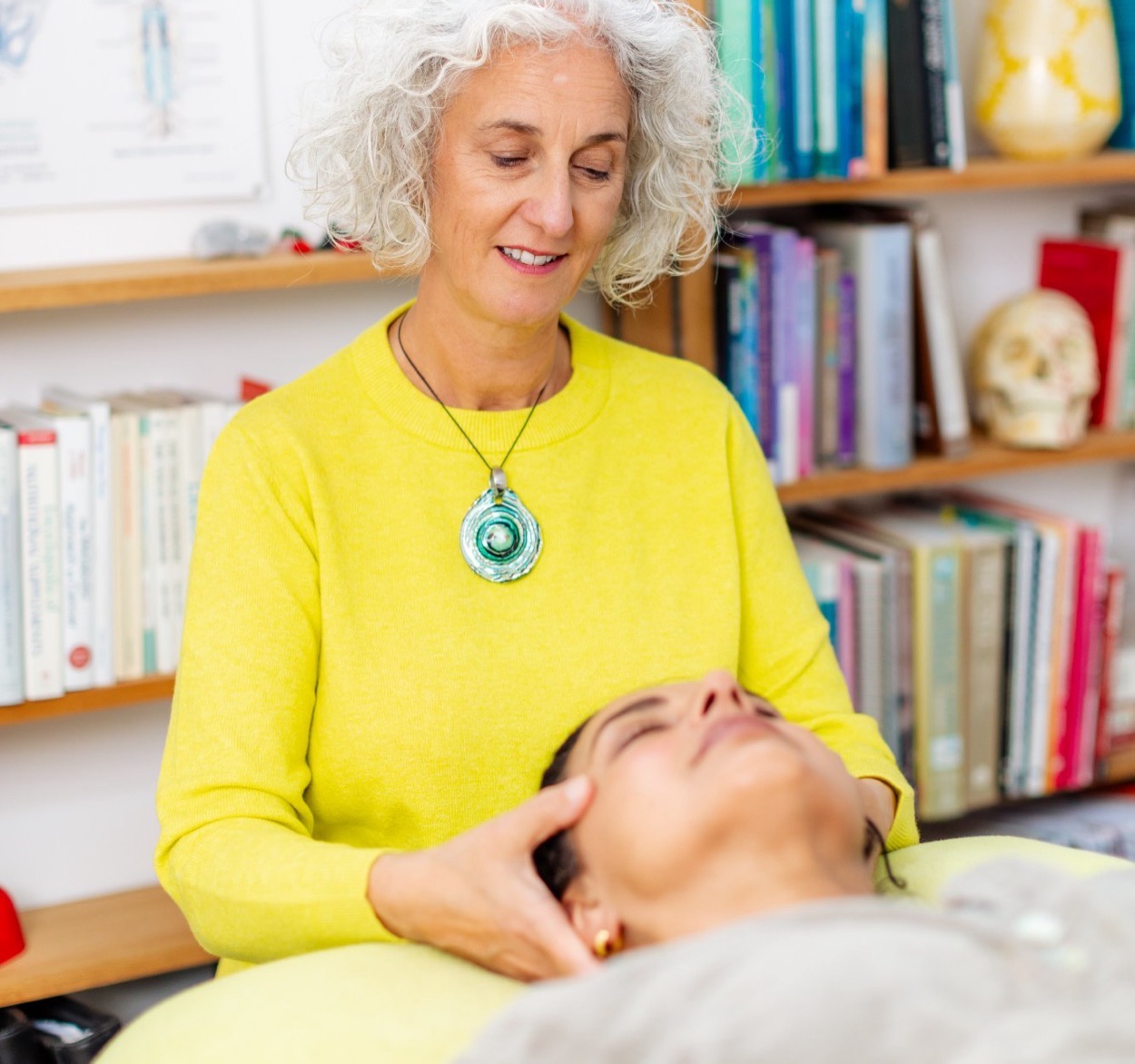 Caroline, in a yellow jumper, smiling as she cradles the head of a woman lying on the treatment table with her eyes closed. Behind them shelves of books, an anatomical chart and a model skull.
