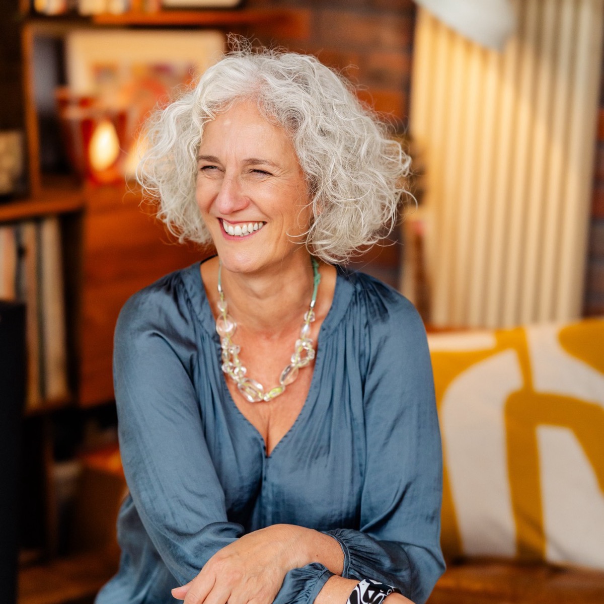 Caroline Barrow, laughing, with curly silver hair and a blue-grey blouse, sitting with her arms folded over one knee. Bookshelves and a warm lamp behind her.